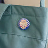 A round pin badge featuring a cartoon cat reading a book, pinned onto a sage green canvas tote bag.