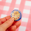 A hand holding a round pin badge featuring a yellow cat reading a book with red and blue details, set against a pink and white checkered background.