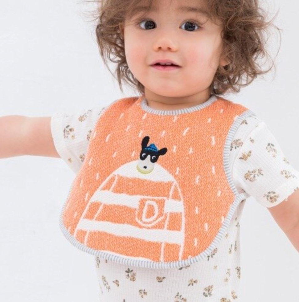 A toddler wearing a textured orange bib with white stripes, a white dog graphic, and a grey trim over a white short-sleeve shirt with a small floral print, set against a plain white background.