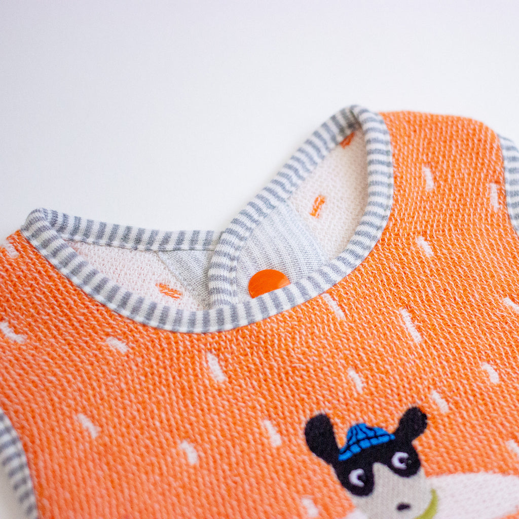 Close-up of a knitted baby bib in bright orange with a grey and white striped binding and a small cartoon animal face.