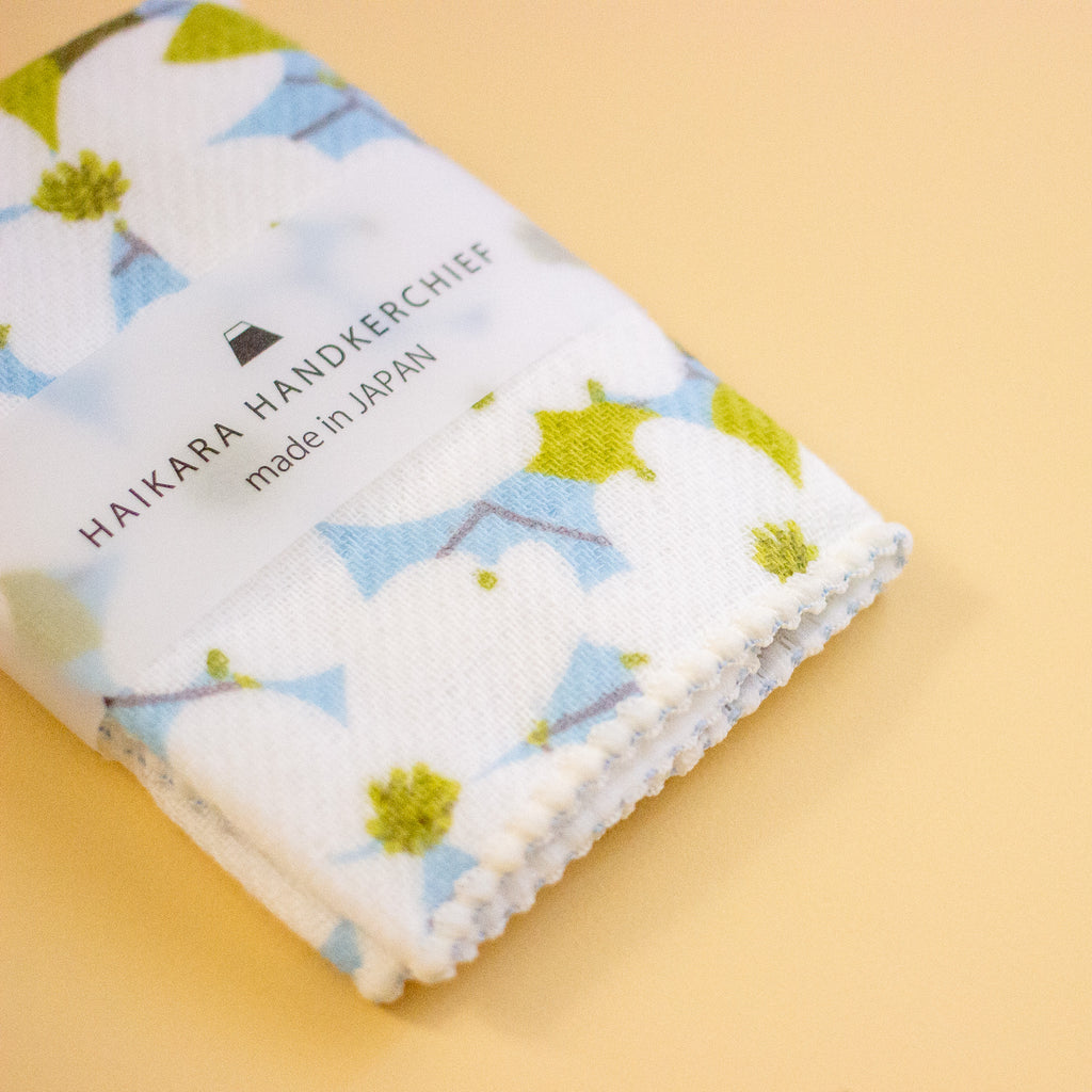 A folded white handkerchief with a light blue and olive green floral pattern, wrapped in a clear paper band that reads 'HAIKARA HANDKERCHIEF made in JAPAN', set against a solid pale yellow background.