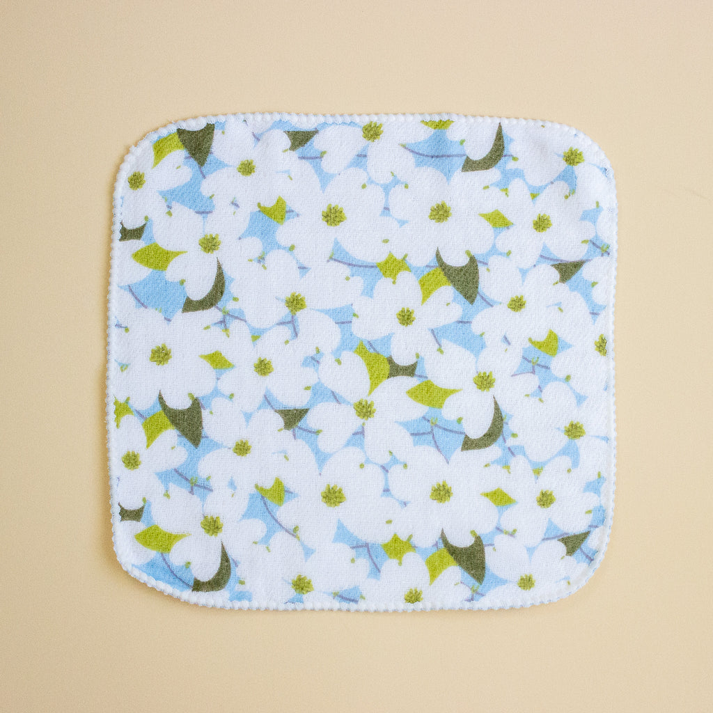 A square handkerchief with rounded corners, featuring a white background printed with a pattern of light blue flowers, white blossoms, and olive green leaves. The cloth has a white merrowed border and is photographed flat against a solid beige background.