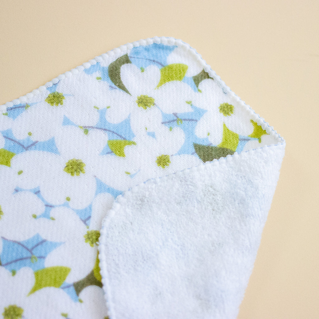 A close-up of a soft white fabric item, featuring a light blue and olive green floral pattern on one side and a plain white textured reverse side, set against a solid beige background.