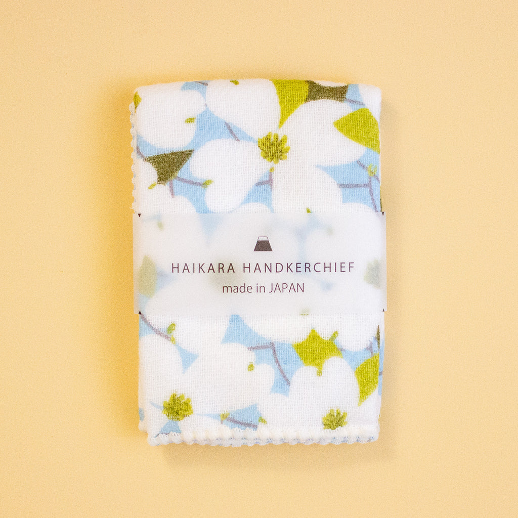 A folded white handkerchief with a light blue and olive green floral print, wrapped in a clear paper band labeled 'HAIKARA HANDKERCHIEF made in JAPAN', set against a solid pale yellow background.