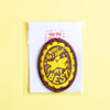 An oval embroidered patch on a white backing card, photographed against a pale yellow background. The patch is brown with a yellow scalloped border and yellow text reading 'WHAT AM I DOING MY BEST'.