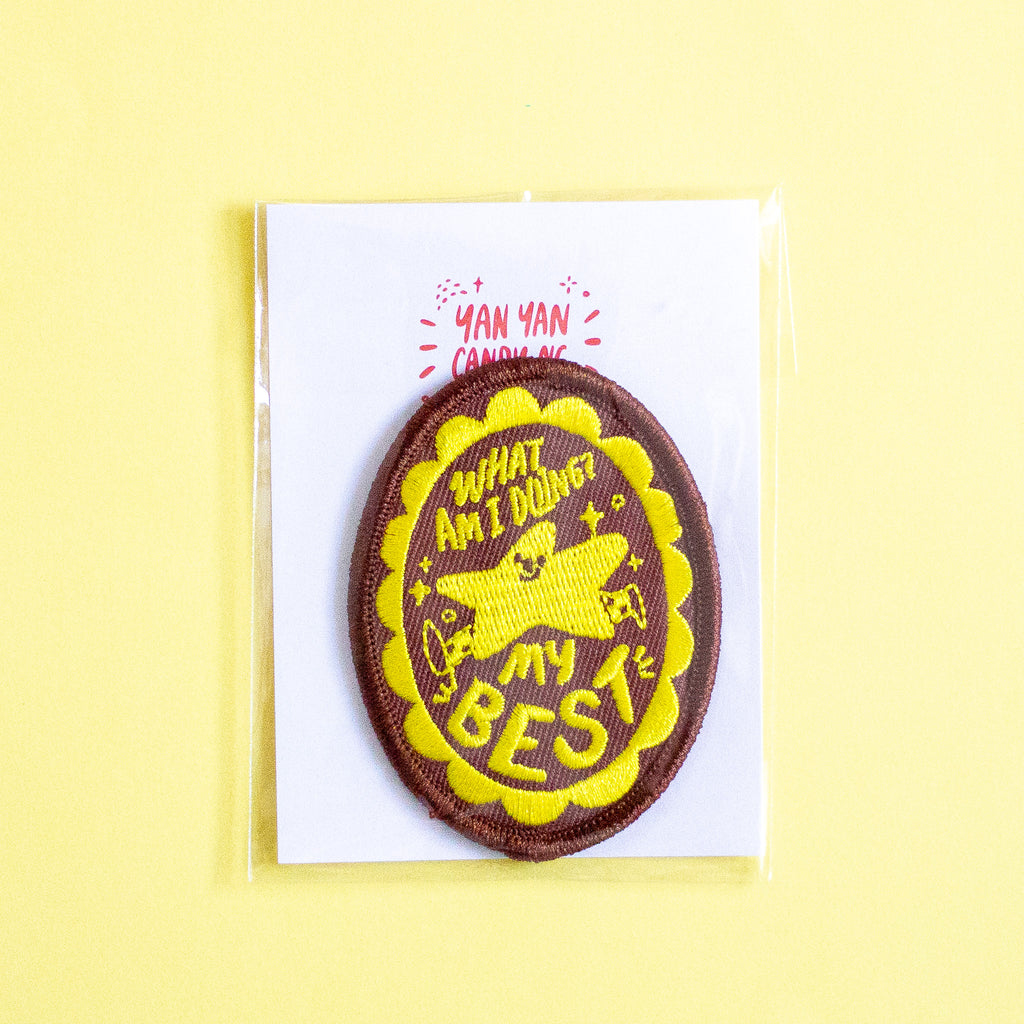 An oval embroidered patch on a white backing card, photographed against a pale yellow background. The patch is brown with a yellow scalloped border and yellow text reading 'WHAT AM I DOING MY BEST'.