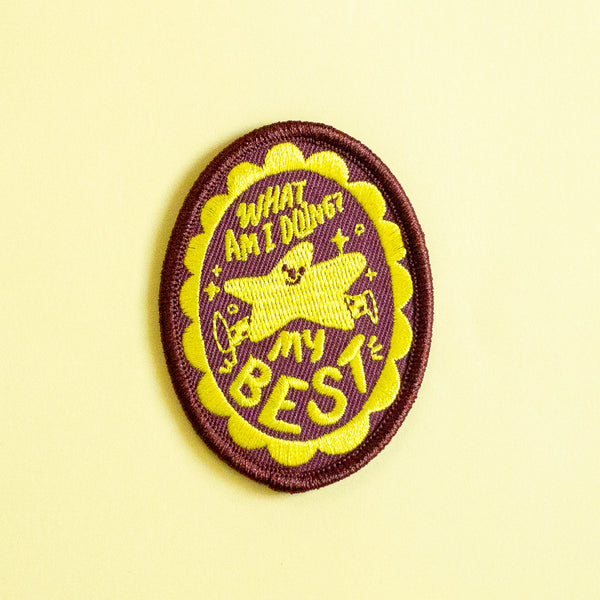 An oval embroidered patch with a dark brown and bright yellow colour scheme, featuring a star character and the text 'WHAT AM I DOING? my BEST'. The patch is displayed on a solid pale yellow background.