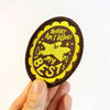 A hand holding an oval embroidered patch with a brown and yellow design that reads 'WHAT AM I DOING MY BEST' with a star illustration, against a plain white background.