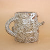 Closeup of the doink figurine, focus on the other side. A large mug shaped sculptural piece made of stoneware clay with natural glaze. Features mouth, eyes, and nose, all hand carved into the figurine. 