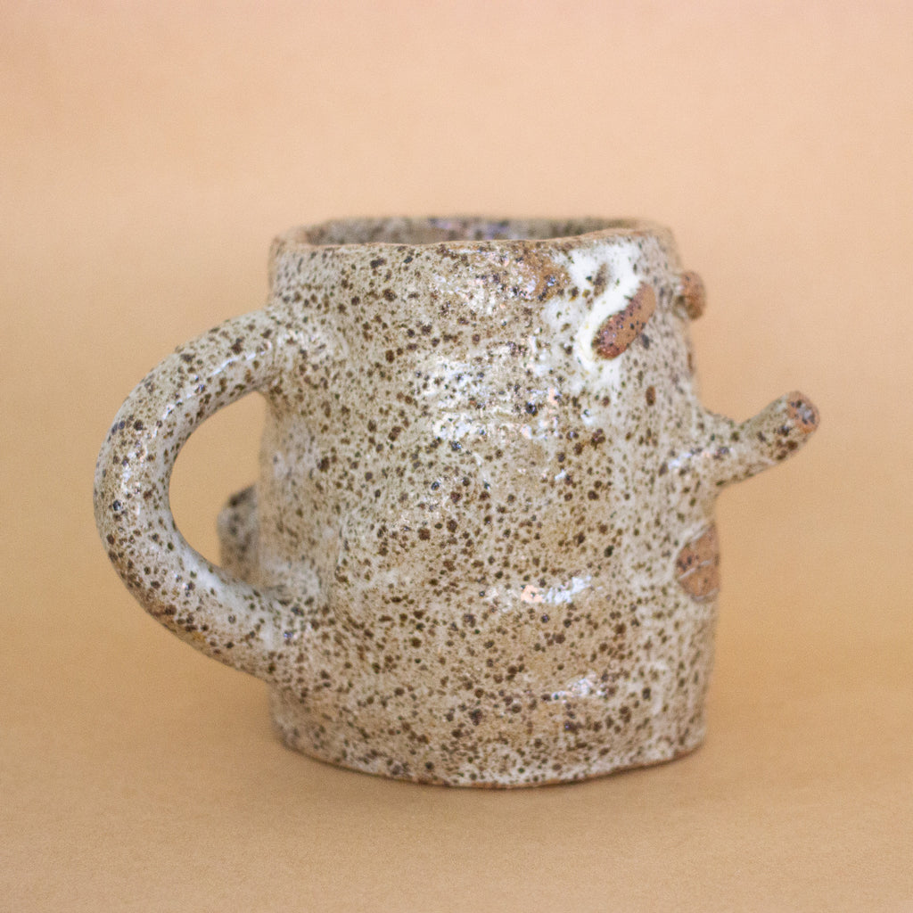 Closeup of the doink figurine, focus on the other side. A large mug shaped sculptural piece made of stoneware clay with natural glaze. Features mouth, eyes, and nose, all hand carved into the figurine. 