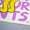 A close-up of a graphic art print featuring bold, stylized lettering in yellow and purple with black outlines on a white paper background, resting on a gray surface.