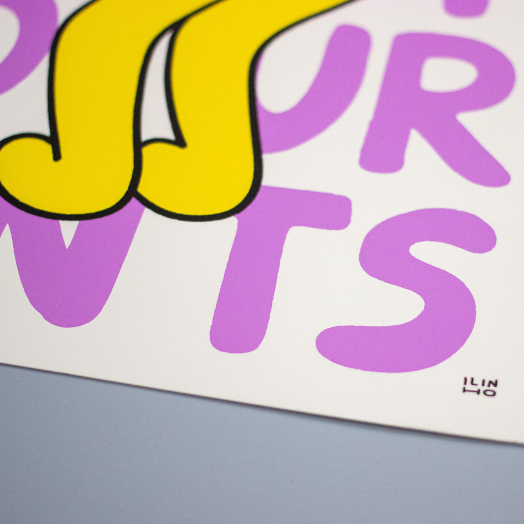 A close-up of a graphic art print featuring bold, stylized lettering in yellow and purple with black outlines on a white paper background, resting on a gray surface.