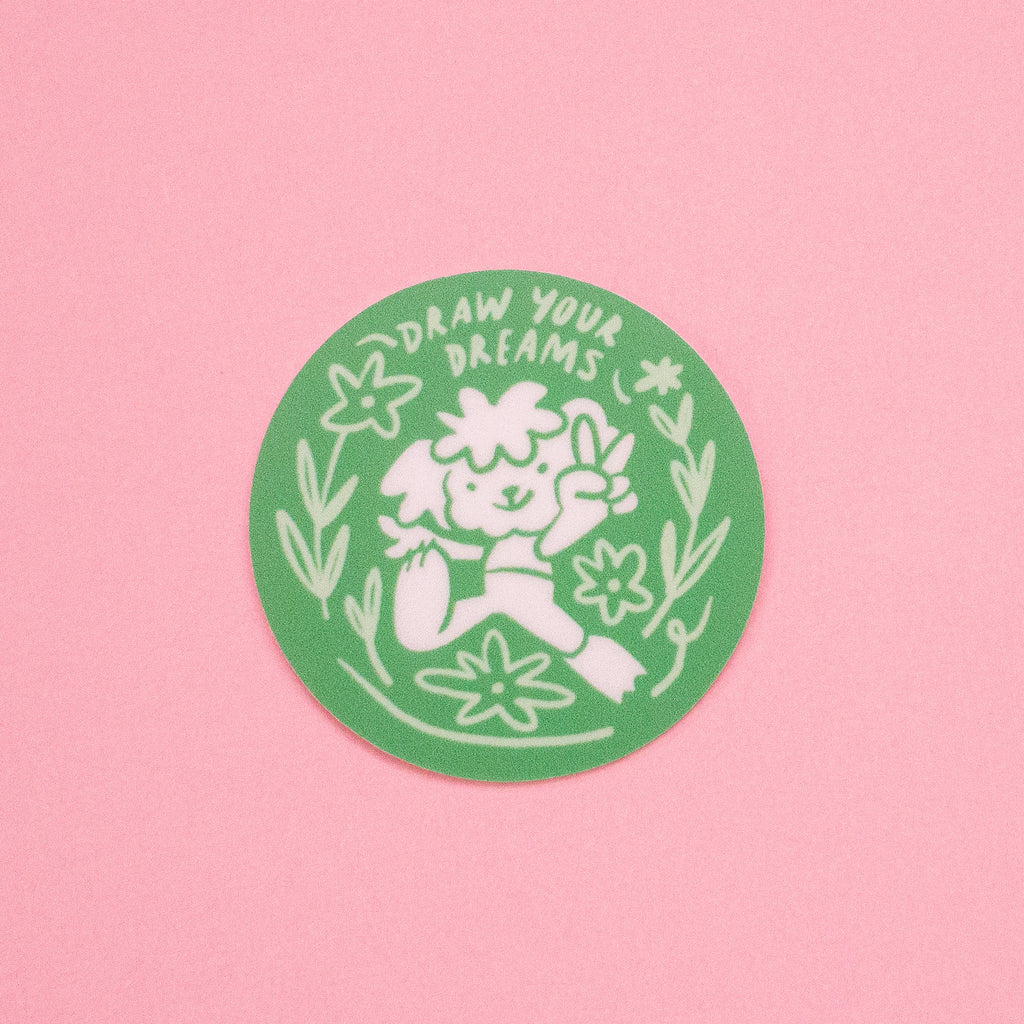 A round green sticker with a white hand-drawn illustration of a cartoon surrounded by flowers and the text 'DRAW YOUR DREAMS', displayed on a solid pink background.