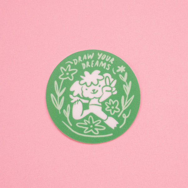 A round green sticker with a white hand-drawn illustration of a cartoon surrounded by flowers and the text 'DRAW YOUR DREAMS', displayed on a solid pink background.