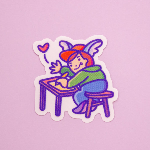 A die-cut sticker featuring a cute cartoon character with wings on a cap, sitting at a desk and writing, on a solid pink background.