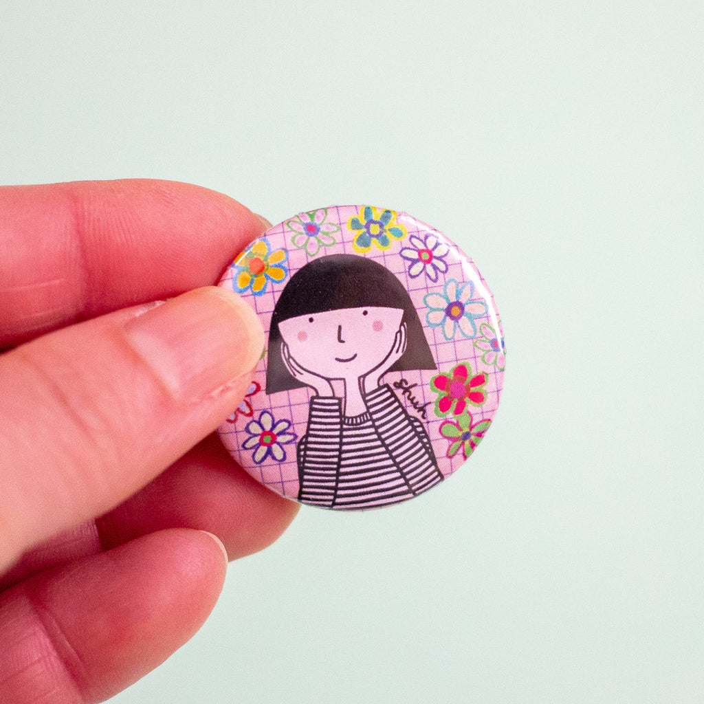 A hand holding a round pinback button badge featuring a cartoon girl with black hair and a striped shirt, surrounded by colourful flowers on a pink grid background, set against a solid pale mint green backdrop.
