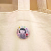 A round pin-back button featuring a cute cartoon girl with black hair and a striped shirt, surrounded by colourful flowers on a pink background, pinned onto a beige canvas tote bag.