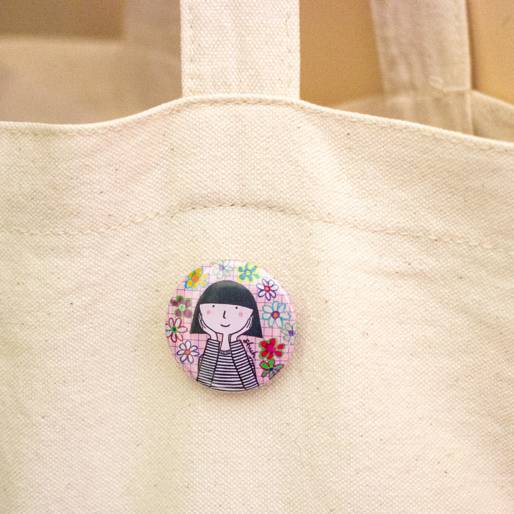 A round pin-back button featuring a cute cartoon girl with black hair and a striped shirt, surrounded by colourful flowers on a pink background, pinned onto a beige canvas tote bag.