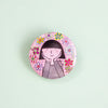 A round pinback button featuring a cartoon illustration of a girl with black hair and a striped shirt, surrounded by colourful flowers on a pink grid background, set against a solid pale mint green backdrop.