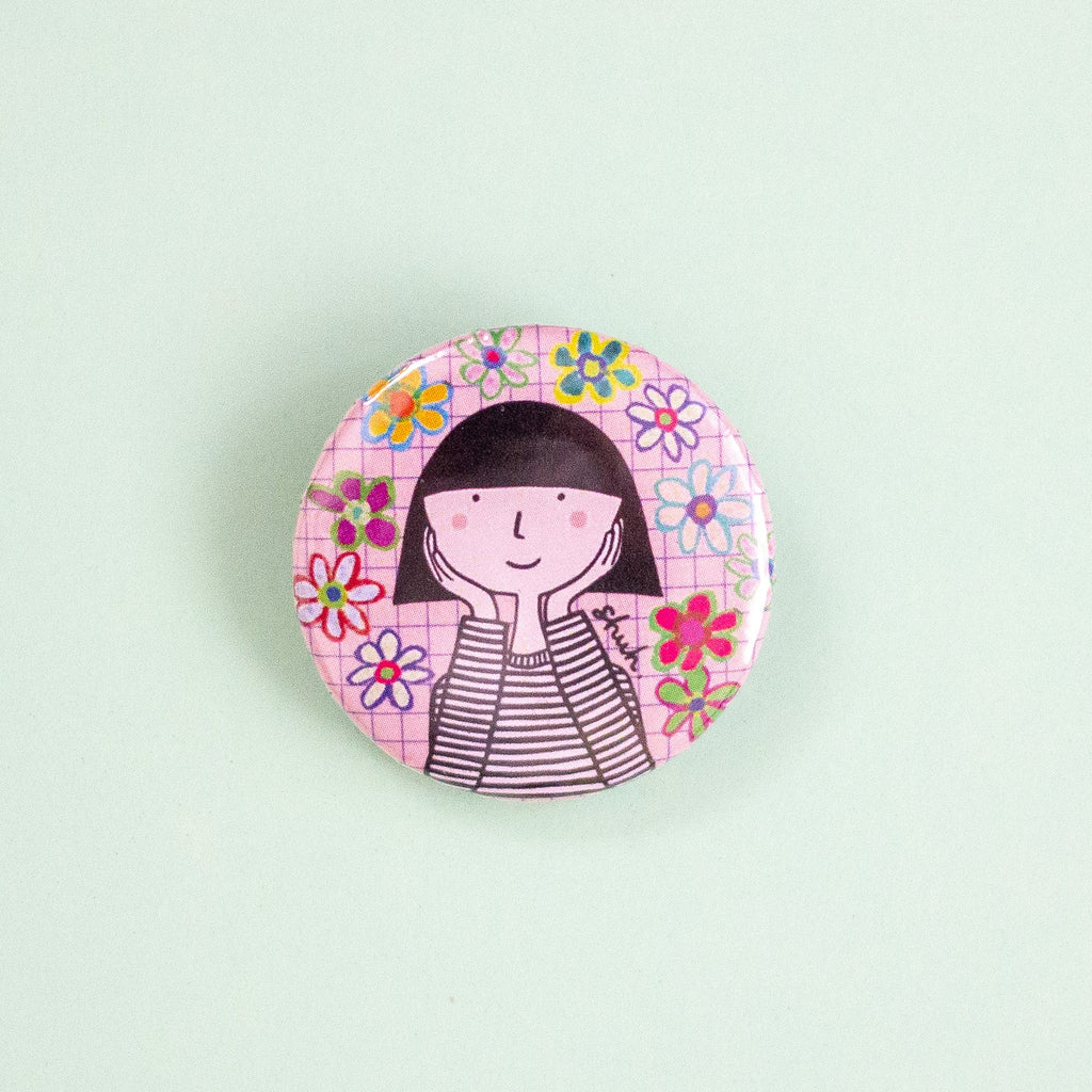 A round pinback button featuring a cartoon illustration of a girl with black hair and a striped shirt, surrounded by colourful flowers on a pink grid background, set against a solid pale mint green backdrop.