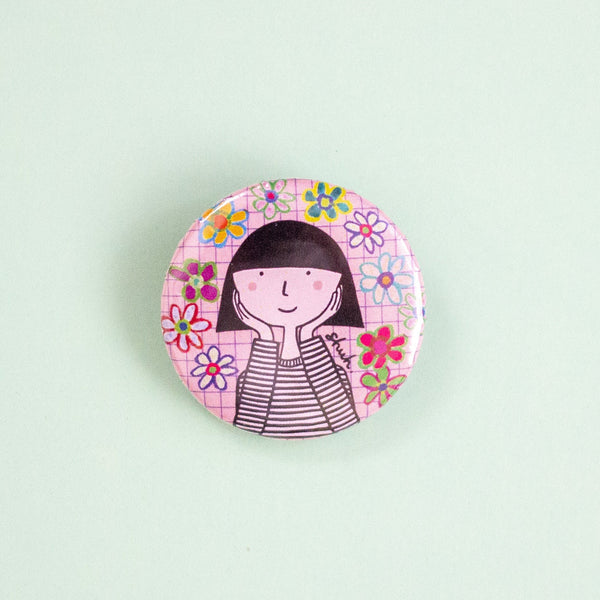 A round pinback button featuring a cartoon illustration of a girl with black hair and a striped shirt, surrounded by colourful flowers on a pink grid background, set against a solid pale mint green backdrop.