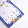 A close-up of a decorative notepad or paper pad with a whimsical illustration. The pad has a white center with gold stars, pink cherries, blue bows, and orange boots, surrounded by a textured dark blue border.