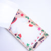 A close-up of a decorative paper notepad with a whimsical illustration of an animal, berries, cherries, flowers, and leaves on a light beige background, photographed on a white surface.