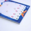 A close-up of a rectangular notepad with a whimsical animal and floral illustration, featuring a white floral dress, gold stars, and orange boots on a dark blue background.