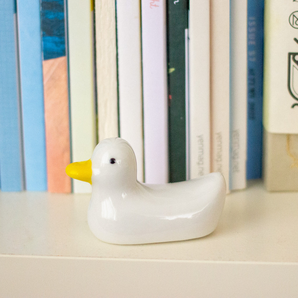 A small glossy white duck figurine with a bright yellow beak, placed on a light surface in front of a row of colourful books.