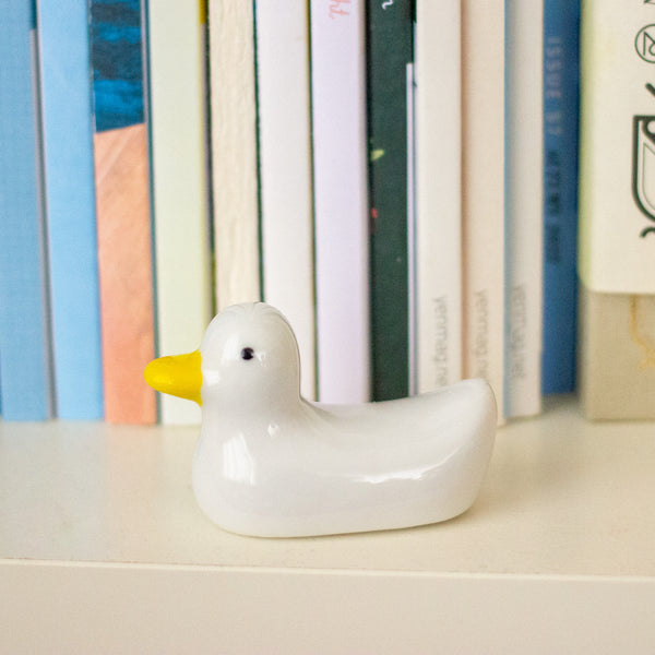 A small glossy white duck figurine with a bright yellow beak, placed on a light surface in front of a row of colourful books.