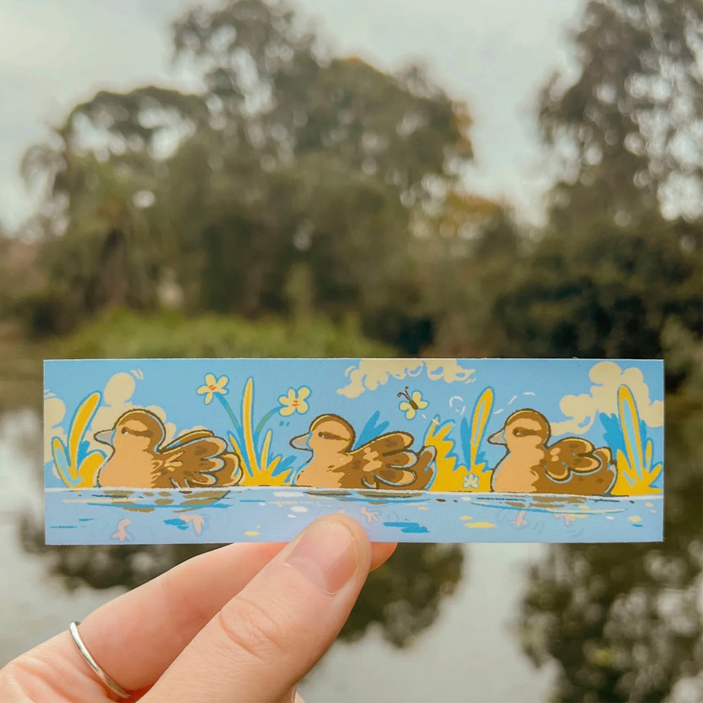 A hand holds a rectangular sticker featuring three cartoon ducklings in a pond with yellow flowers and a blue sky background, with a blurred outdoor pond and trees behind it.
