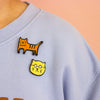 Light blue shirt with cat-themed patches on a peach background