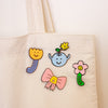 An off-white canvas tote bag with four colourful enamel pins attached to the front. The pins feature cute cartoon designs: a yellow smiling flower with blue stems, a light blue smiling vase with a yellow flower, a pink smiling flower, and a pink bow with a yellow smiley face center.