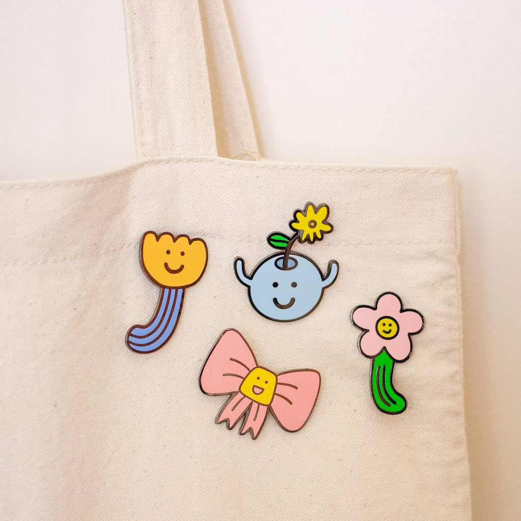 An off-white canvas tote bag with four colourful enamel pins attached to the front. The pins feature cute cartoon designs: a yellow smiling flower with blue stems, a light blue smiling vase with a yellow flower, a pink smiling flower, and a pink bow with a yellow smiley face center.
