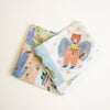 Two folded fabric items with whimsical illustrated prints, including a fairy holding a flower and a landscape scene, displayed on a light cream background.