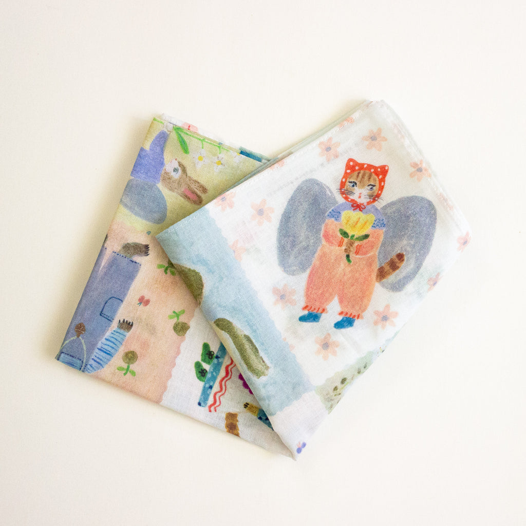 Two folded fabric items with whimsical illustrated prints, including a fairy holding a flower and a landscape scene, displayed on a light cream background.