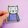 A hand holding a die-cut sticker featuring a cute cat character wearing a blue and white stripe top with a holographic border, set against a plain light purple background.