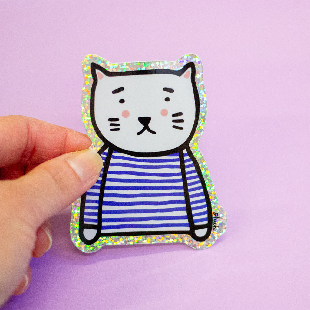 A hand holding a die-cut sticker featuring a cute cat character wearing a blue and white stripe top with a holographic border, set against a plain light purple background.