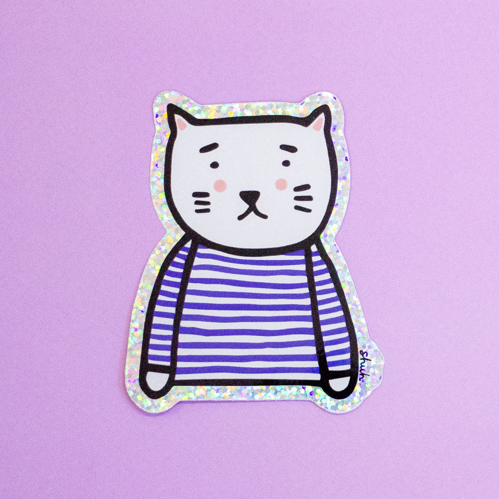 A die-cut sticker featuring a cute cat character wearing a blue and white stripe top with a holographic border, set against a plain light purple background.