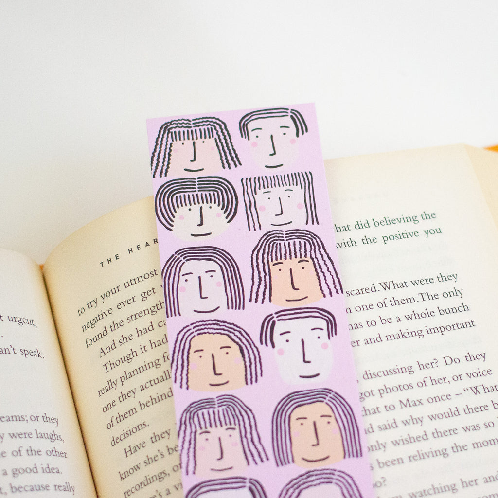 A pink bookmark with minimalist line-art illustrations of faces with long hair, placed on open book pages.
