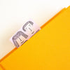 A close-up of a bright yellow textured notebook or folder with a small pink bookmark or tag peeking out from the top, printed with a simple black line drawing of two faces.