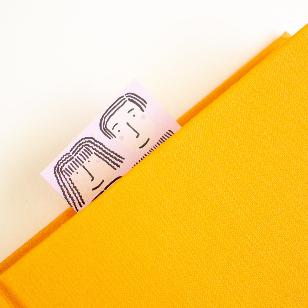 A close-up of a bright yellow textured notebook or folder with a small pink bookmark or tag peeking out from the top, printed with a simple black line drawing of two faces.