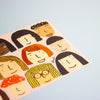 A close-up of a printed illustration sheet featuring multiple stylised cartoon faces in warm tones on a pale blue background.