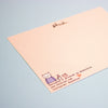 A pink-coloured postcard with a small printed illustration of a cat and text at the bottom, photographed on a light blue background.