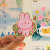 A hand holds a die-cut sticker featuring a pink bunny character with glittery green wings and a small star wand, set against a colourful, softly blurred background of posters and stationery.