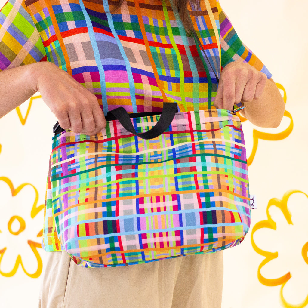 Farm easy bag used as a shoulder bag by model. Print is bright coloured lines in a grid pattern. 