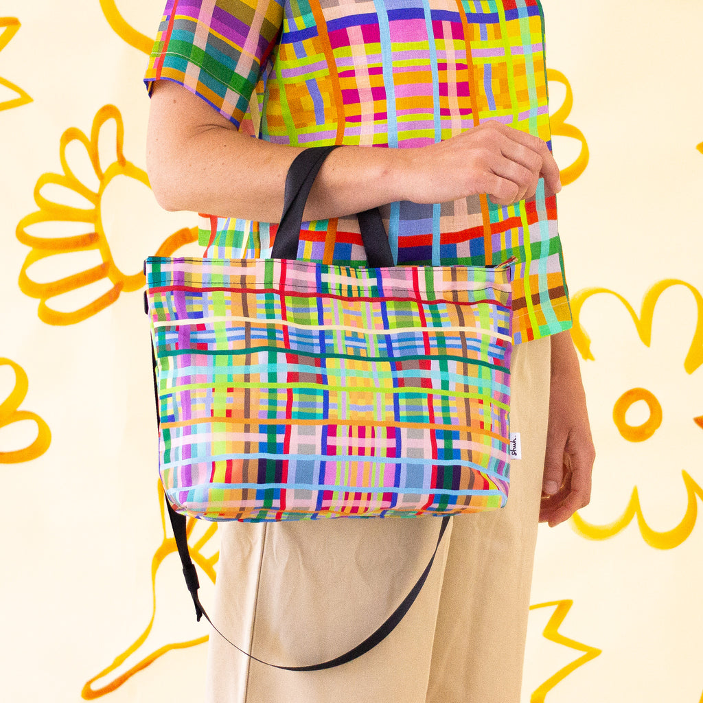 Farm easy bag used as a tote bag by model. Print is bright coloured lines in a grid pattern. 