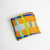 A tote bag folded into a square with a vibrant multicolour plaid pattern, featuring orange, blue, green, yellow, and pink blocks, photographed on a clean white background.