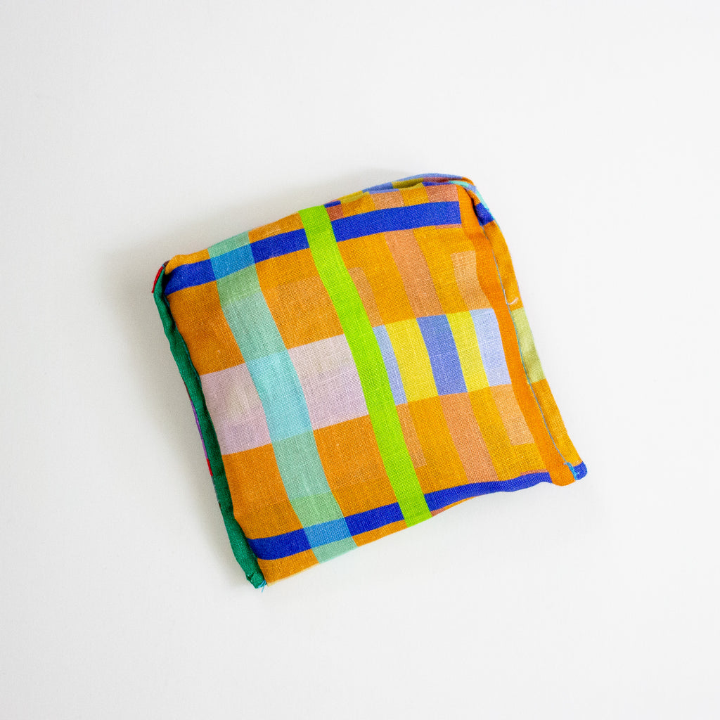 A tote bag folded into a square with a vibrant multicolour plaid pattern, featuring orange, blue, green, yellow, and pink blocks, photographed on a clean white background.