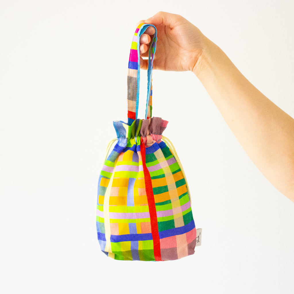 Front view of the Farm mini carry bag held by a hand. Multicoloured stripes, giving an impressionist birds eye view of fields. 